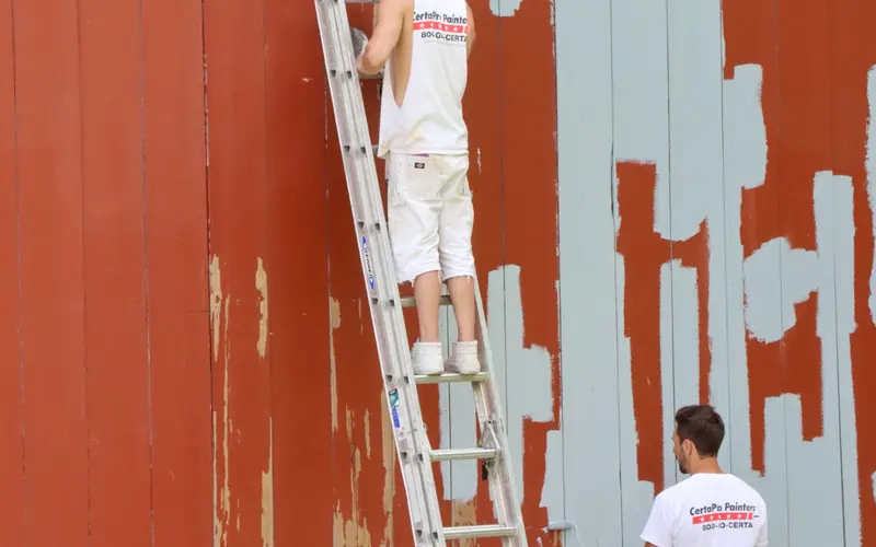 Two painters on a ladder repainting a red wooden house wall in a light blue-gray color